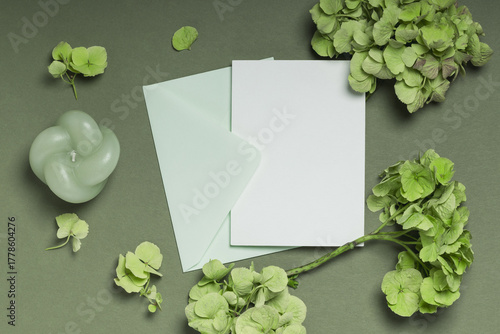 Blank card and envelope with greenish hydrangea branches and handmade candles on a green background. Invitation or greeting card concept.