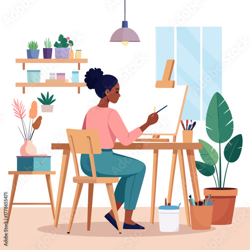 A focused Black woman artist in a pink shirt and teal pants painting on a canvas at her desk.