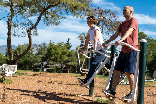 A middle aged Caucasian couple is exercising  using outdoor fitness equipment outdoors at a public park.