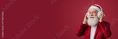 Man dressed as santa claus wearing red headphones and listening to music with eyes closed. Smiling elderly man in red suit enjoying holiday tunes. Christmas and new year entertainment concept for