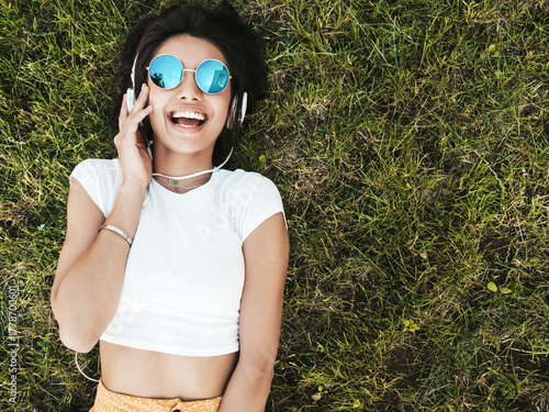 Fashion portrait of young stylish hipster woman lying on the grass in the park.She weares trendy outfit.Smiling model enjoy her weekends. Female listening to music via headphones.Top view