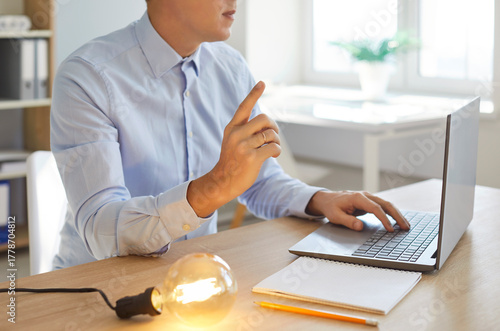Young intelligent businessman with laptop, paper and glowing light bulb changes marketing plan using genius brain, knowledge and new smart ideas. Business creativity, technology, inspiration concept © Studio Romantic