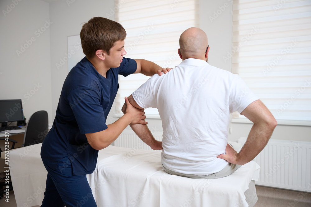 Obraz premium Chiropractor adjusting male patient’s shoulder in clinic. Physiotherapist performing manual therapy to relieve pain, improve mobility, support rehabilitation after injury or musculoskeletal disorder.