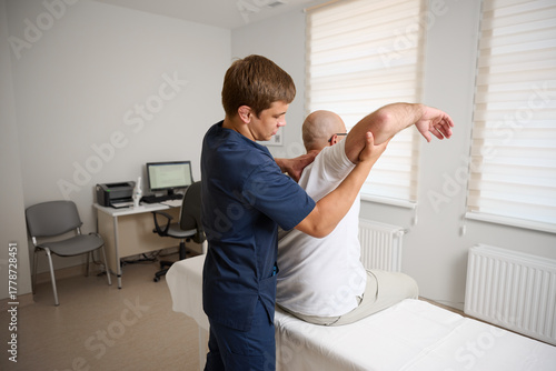 Chiropractor adjusting male patient’s shoulder in clinic. Physiotherapist performing manual therapy to relieve pain, improve mobility, support rehabilitation after injury or musculoskeletal disorder.
