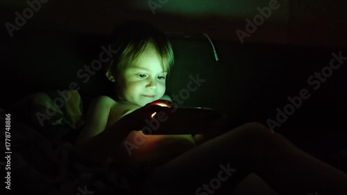 A little boy is playing mobile games on her phone. Addiction to online video games on a smartphone. A preschooler child plays on the phone at night