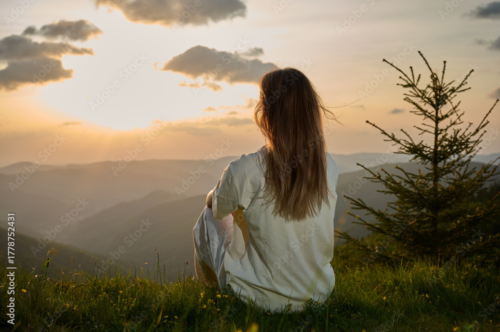 Obraz premium Woman sits peacefully on a grassy hill, gazing at the sunset as golden light bathes the landscape. Rolling mountains and a single evergreen tree create a tranquil atmosphere in the evening.