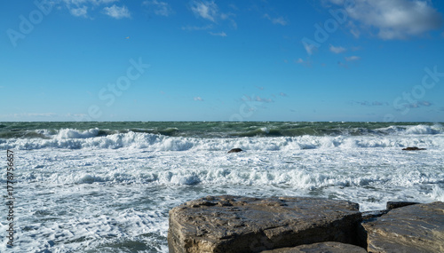 Coastal seascape with large flat rocks in the foreground and turbulent sea waves crashing onto the shore. The sky is clear with scattered clouds. Bright sunlight highlights the texture of wet stone.