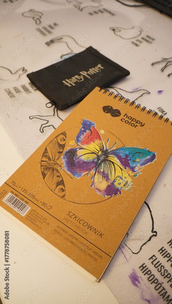 Naklejka premium A 'Happy Color' drawing notebook adorned with a vibrant butterfly illustration rests on a table alongside a 'Harry Potter' branded pouch, hinting at a casual moment of artistic creativity