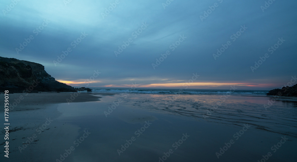 Obraz premium The serene beach at sunset features gentle waves kissing the sandy shore, surrounded by rugged cliffs on Portugal's coastline. The sky's pastel hues add a calming ambiance, perfect for a tranquil.