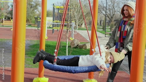 Mom and her daughter swinging on swing in autumn park. Happy family, child outdoors, weekend. Child girl, sits on swing smiles. Playground in fall. 