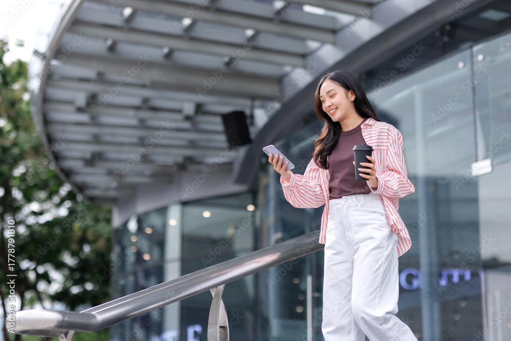Fototapeta premium Happy young asian woman walking, using smartphone, holding coffee