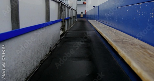 Showing empty walkway extending along bench inside ice hockey rink, with blue handrail and clock
