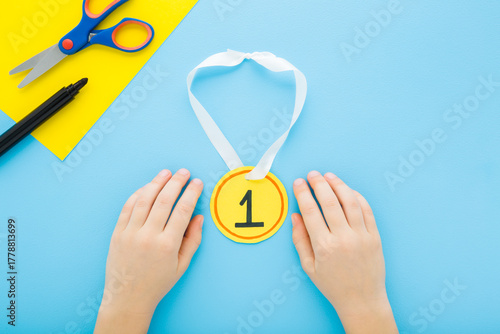 Little child boy hands making and creating yellow gold paper medal with white ribbon on light blue table background. Pastel color. Closeup. Point of view shot. Flat lay. Top down view.
