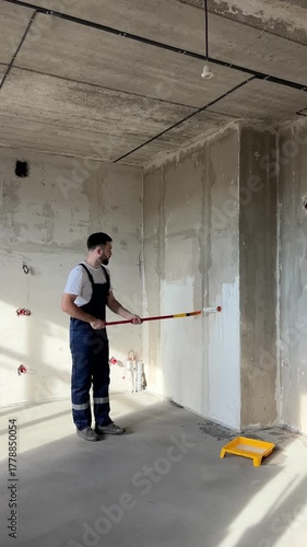 A bearded man in a work uniform is priming the walls after applying gypsum plaster. 