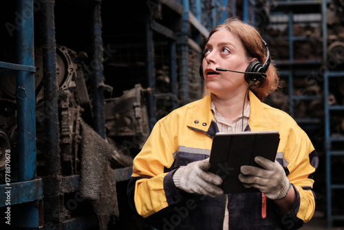 One Hispanic mature female inspector remotely works with headset and tablet, checks auto spare parts stock in warehouse stack, operator jobs in manufacturing factory, and industrial worker occupation.