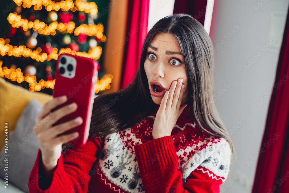 Naklejka premium Funny surprise as a cheerful woman in a red christmas sweater chats on her phone at home with a glowing christmas tree in the background