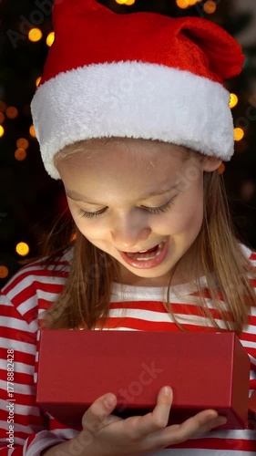 Happy girl in red santa hat opens gift box with magic light and smiles on background of christmas tree. New Years present. Vertical shot
