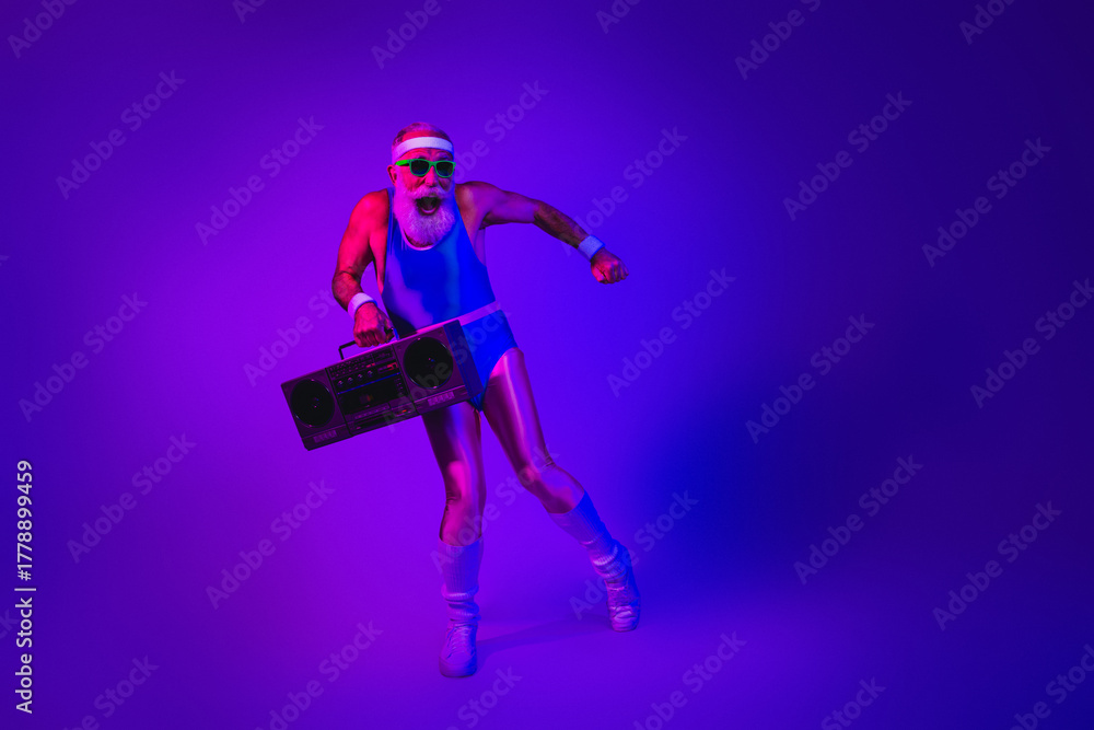Fototapeta premium Energetic bearded senior in retro fitness outfit dancing with a boombox under neon purple and blue lighting