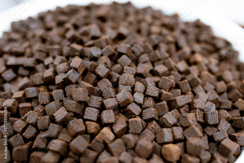 A close-up macro view of small, brown, textured pellets. These compressed fibrous cubes could be biomass fuel, animal feed, a food ingredient, or a nutritional supplement.