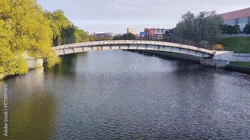 A curved, modern bicycle bridge spans across a river, surrounded by lush greenery. The bridge with railings on both sides. Video 