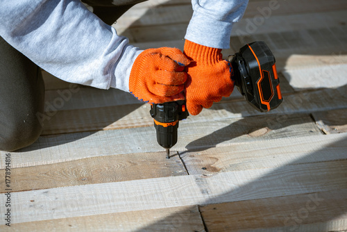 Φωτογραφία A close-up of a worker's hands in orange gloves using a cordless electric drill to drive a screw into a new wooden deck