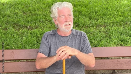 Happy cheerful elder 70s man with walking disability holding knob of cane, sitting on a bench in the park, suffering from muscles problems, bones disease, recovering after trauma