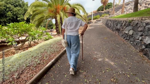 Back view of elder 70s woman with walking disability holding knob of cane, leaning on gimp stick walking in the park, suffering from muscles problems, bones disease, recovering after trauma