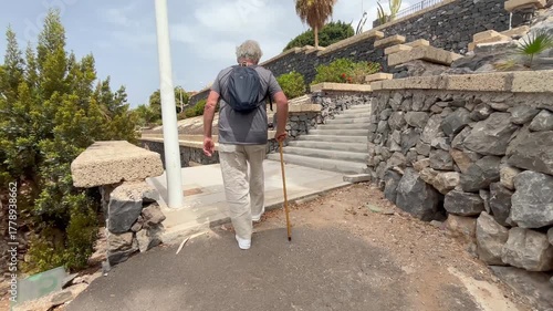 Back view of elder 70s man with walking disability holding knob of cane, leaning on gimp stick walking in the park, suffering from muscles problems, bones disease, recovering after trauma