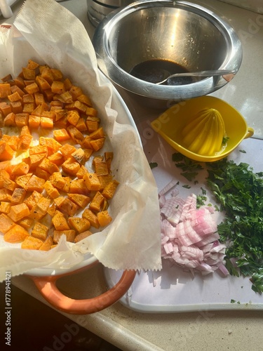 Chopped roasted sweet potatoes rest in a bowl beside chopped onions and herbs, with a lemon juicer and dressing waiting to be mixed for a vibrant salad.