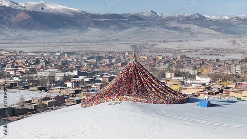 Aerial view of prayer flags cascading down a snowy hill with the city of Litang and snow capped mountains in the background, Litang, China.