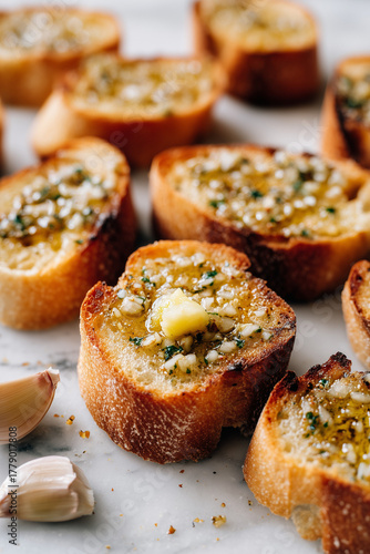 Hot toasted baguette slices, some visibly golden-brown, with a fresh garlic clove being rubbed over the surface; the topping sits just aside ready for assembly. vertical image