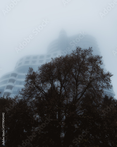 A dramatic, atmospheric shot of a modern multi-story building with a flowing, curved facade and horizontal rows of balconies, which is almost completely hidden in thick fog or haze.
