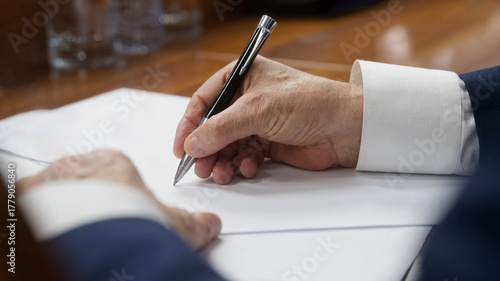 Close-up of an elderly businessman’s hand in a suit writing with a metal pen on blank papers at a wooden desk; legal document or contract moment in a boardroom. Phot