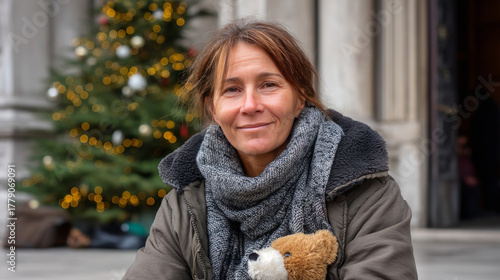 Fototapeta Naklejka Na Ścianę i Meble -  A homeless woman holding a stuffed animal, sitting on the cold ground with a small Christmas tree next to her, wishing for warmth and compassion. As a result of winter's cold, harshness, warmth, holi