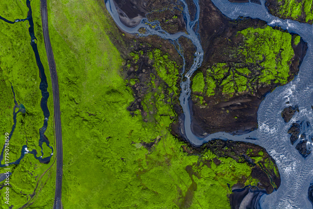 Fototapeta premium Aerial view of neon green moss on dark lava with braided streams near Vik, Iceland. A narrow road lines the left, rivers merge right in crisp daylight contrast.