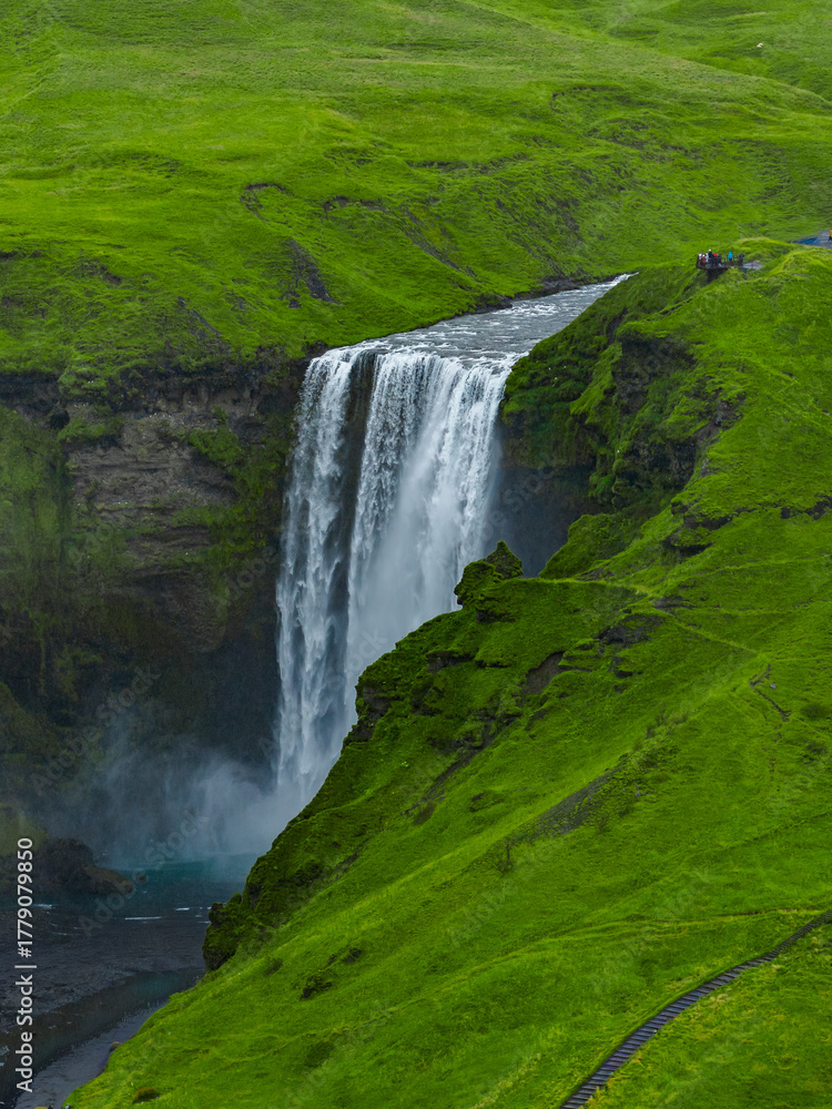 Fototapeta premium A broad Skogafoss waterfall drops over a rocky cliff into a misty gorge in southern Iceland. Mossy hills, winding footpaths, and tiny hikers show scale in soft overcast light.