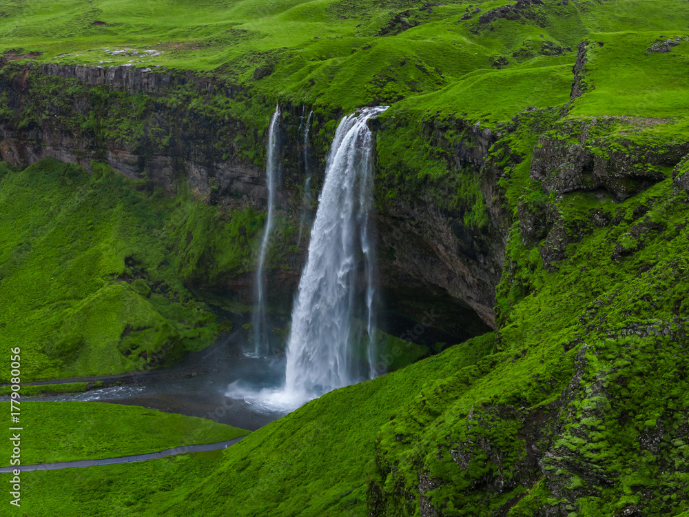 Fototapeta premium Aerial view shows Seljalandsfoss on Iceland South Coast, white water drops over mossy basalt into a shallow pool, footpath and recess behind the falls are visible in soft light.