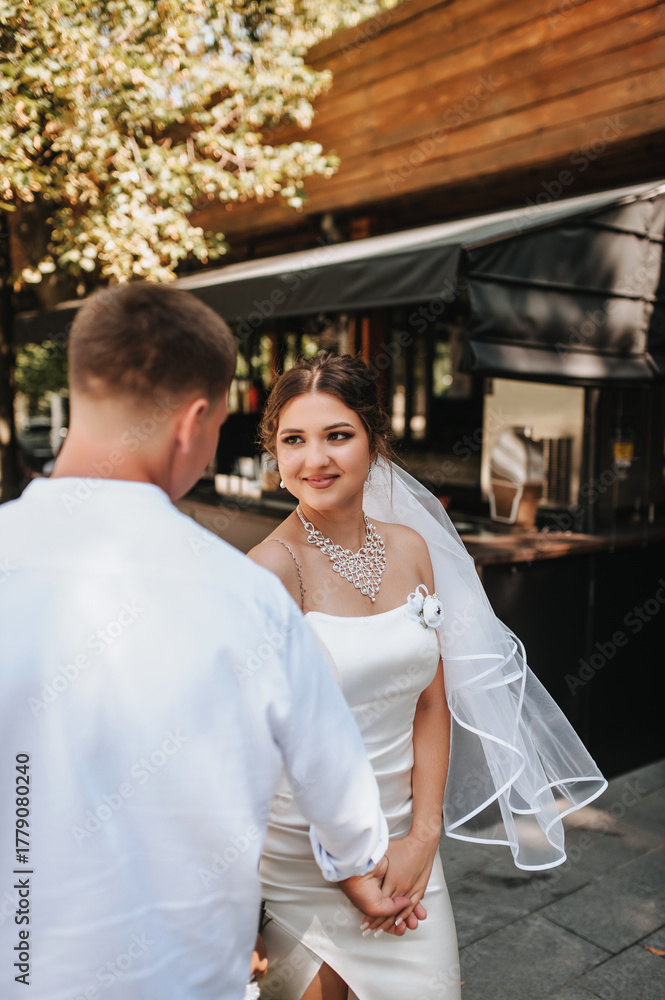 Naklejka premium Wedding photography, portrait of happy smiling newlyweds, stylish groom and beautiful young brunette bride, walking and holding hands, outdoors on the street in the city.
