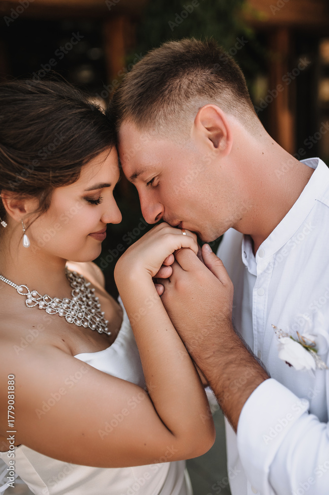 Fototapeta premium Wedding photography, close-up portrait of happy fashionable newlyweds, young stylish groom kissing hand of beautiful brunette bride outdoors.
