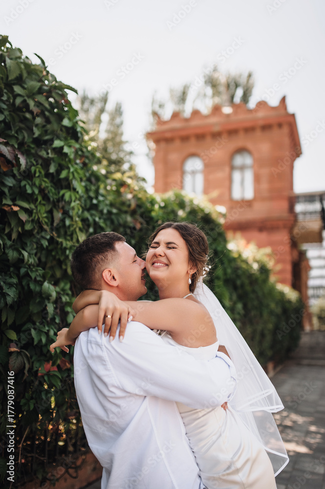 Naklejka premium Wedding photography, portrait of happy smiling newlyweds, a young groom and a beautiful sweet brunette bride, tenderly hugging outdoors on a street in the city, in a park.