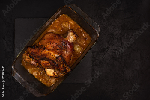 Baked pork knuckle in marinade in a glass bowl on a stone cutting board against a black textured background. Photo of prepared food. Top view with copy space. Cooked in the oven or in the stove