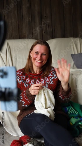 Smiling woman in a cozy sweater waves at her phone camera while knitting and streaming from home. Sharing warmth, creativity, and comfort through a personal video call or vlog.