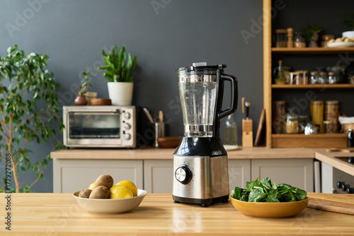 Electric blender standing on kitchen counter surrounded by bowl of fresh spinach leaves and bowl of whole fruits including kiwis and lemons, modern kitchen appliances in background