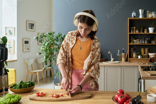 Quadro em tela Teenage girl wearing headphones slicing tomatoes on wooden cutting board in mode