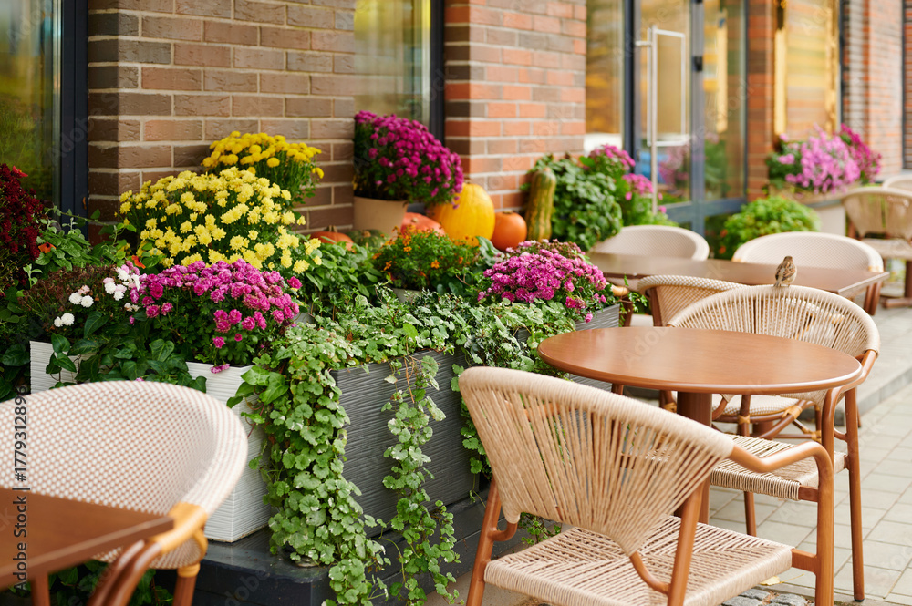 Fototapeta premium outdoor cafe scene with empty wicker chairs and wooden tables surrounded by vibrant flowers and foliage. charming seating arrangement evokes cozy, relaxed ambiance in urban setting