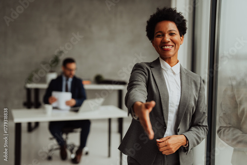 African American businesswoman gesturing while talking in modern office, with male colleague working at desk in background.
