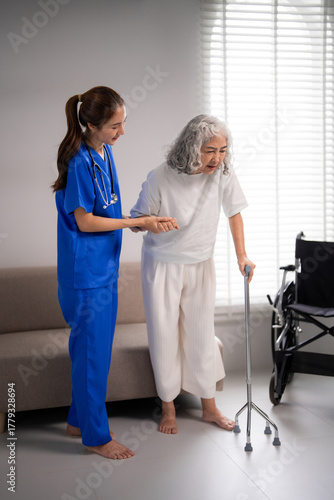 Young Asian adult nurse providing caring assistance older Asian adult woman walking with quad cane home healthcare physical rehabilitation living room setting