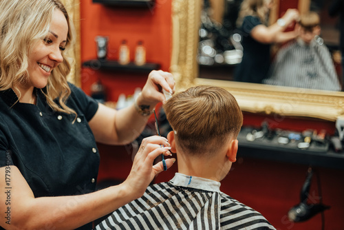 Female hairdresser carefully cuts a young boy's hair in a stylish barbershop with red walls and gold-framed mirrors
