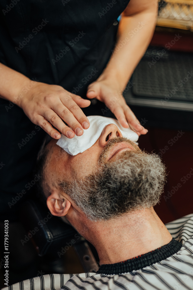 Fototapeta premium Bearded man relaxes in a barber chair while a hot towel is gently applied to his face as part of a grooming or skincare treatment