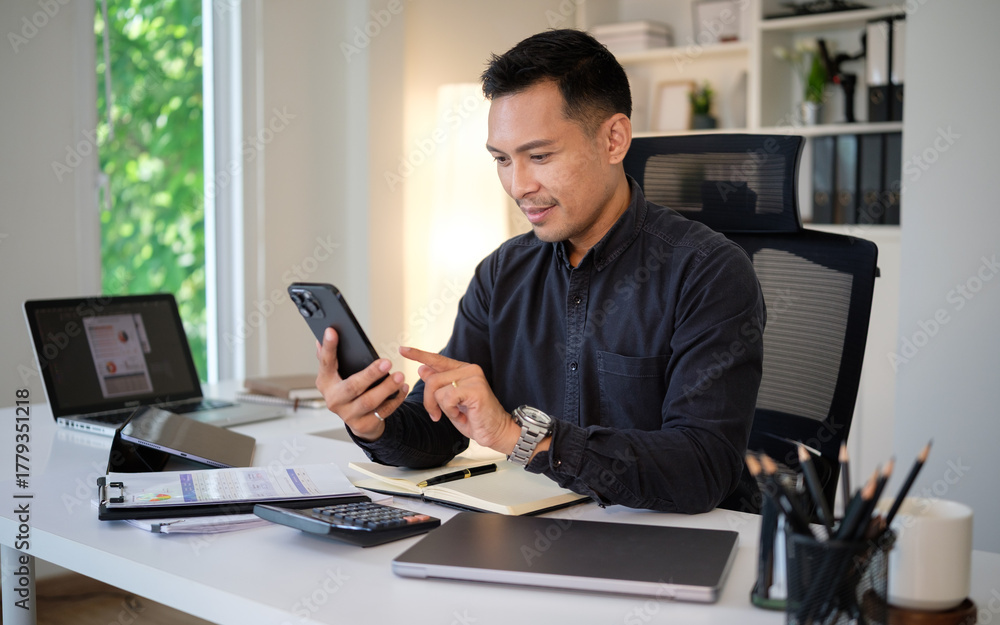 Fototapeta premium Confident businessman using smartphone at modern office desk with laptop and documents.